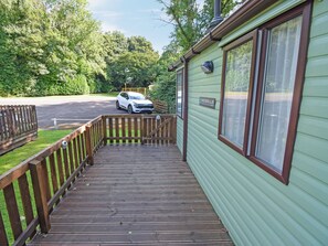 Eine Holzterrasse führt zu einem grünen Gebäude mit einer Namensschild, im Hintergrund ein geparktes Fahrzeug und ein eingezäuntes Gelände.