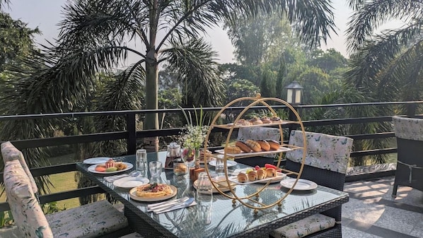 An outdoor dining area with a glass table, wicker chairs, and a tiered bread display. - The Golden Tusk (Ramnagar)