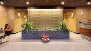 A reception area with a marble counter, two potted plants, a decorative water feature, and a statue of a monk.