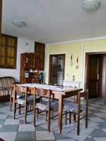 A dining area with a wooden table and chairs, a kitchen in the background, and a yellow wall.