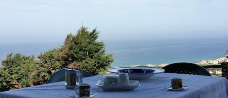 Une table dressée pour le dîner, avec vue sur la mer, un ciel dégagé et quelques plantes vertes en arrière-plan.