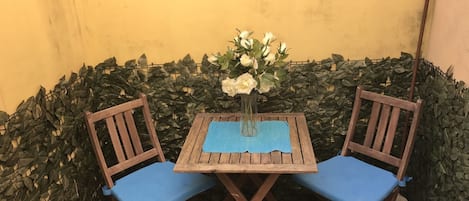 A small wooden table with a vase of white flowers, two wooden chairs with blue cushions, and a yellow wall with green foliage.