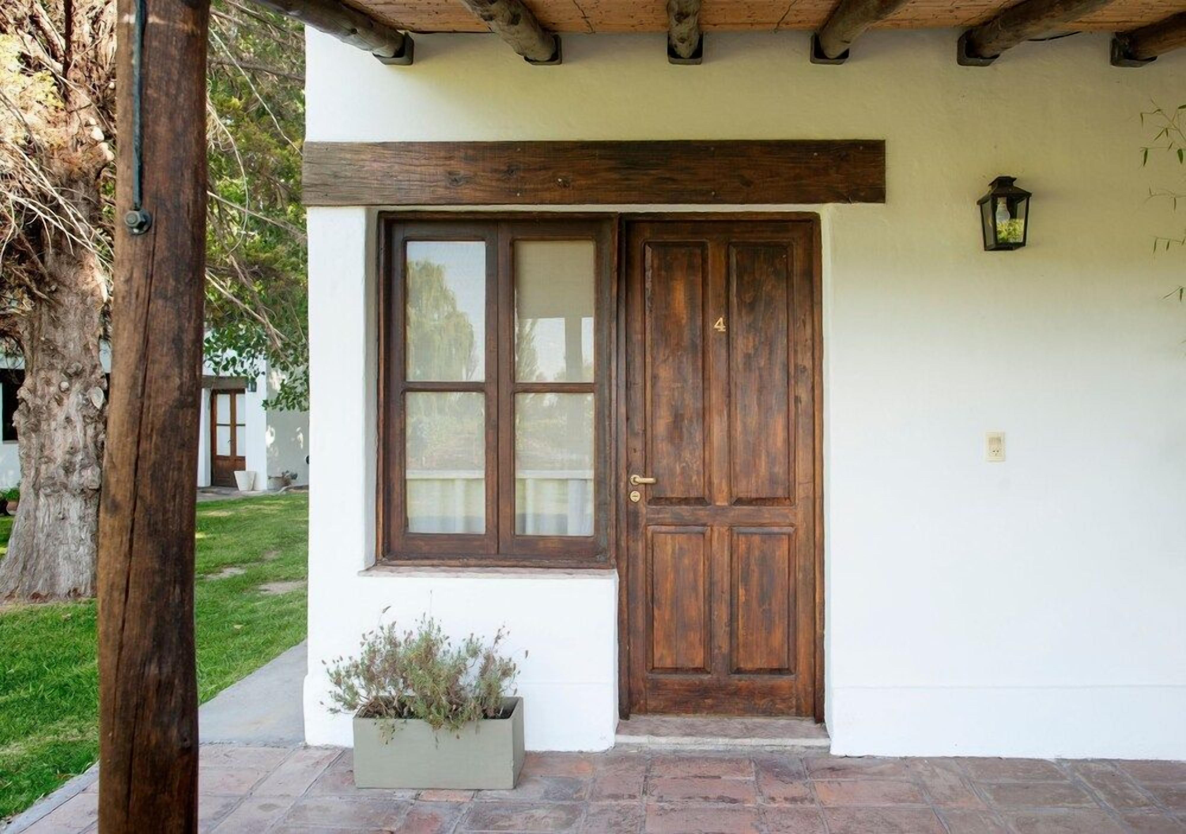 Un edificio blanco con puerta y ventana de madera, una maceta con plantas y un farol en la pared.