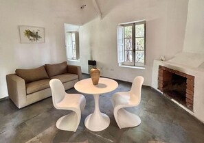 Una sala de estar con un sofá marrón, dos sillas blancas, una mesa redonda, una chimenea y una ventana.