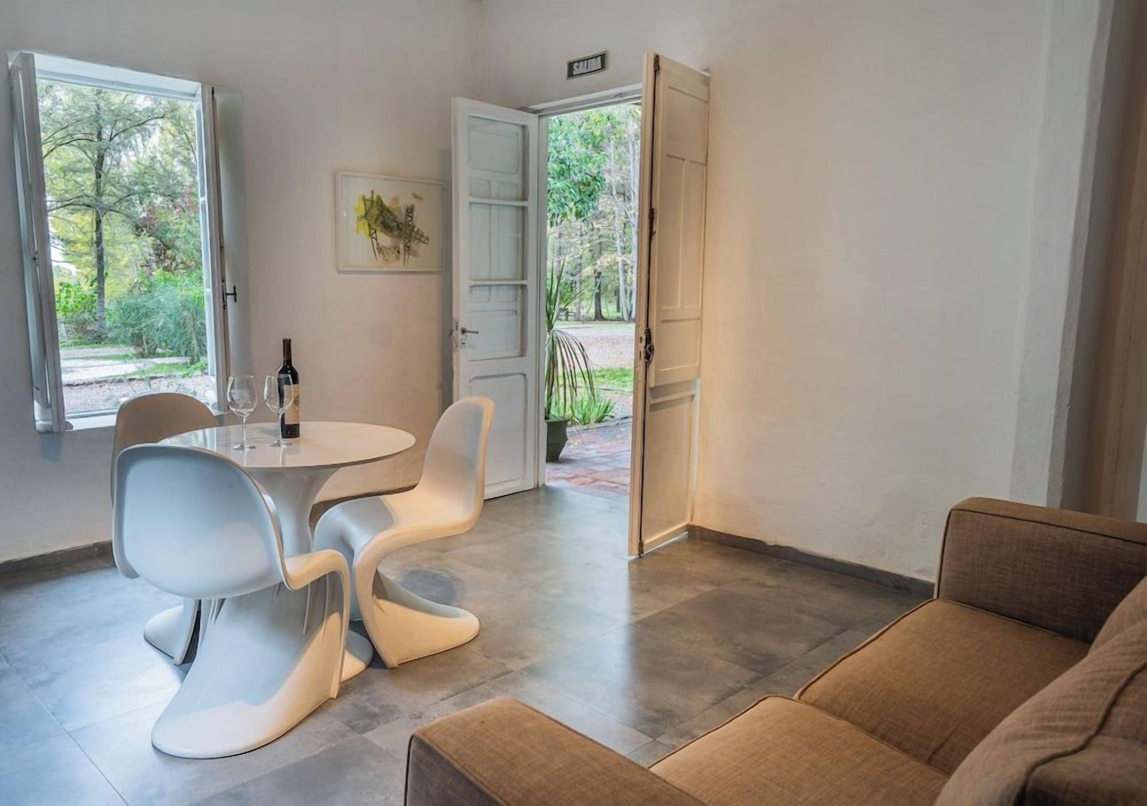 A modern interior with a dining area featuring a round table and white chairs, a sofa, and a door leading to an outdoor garden.