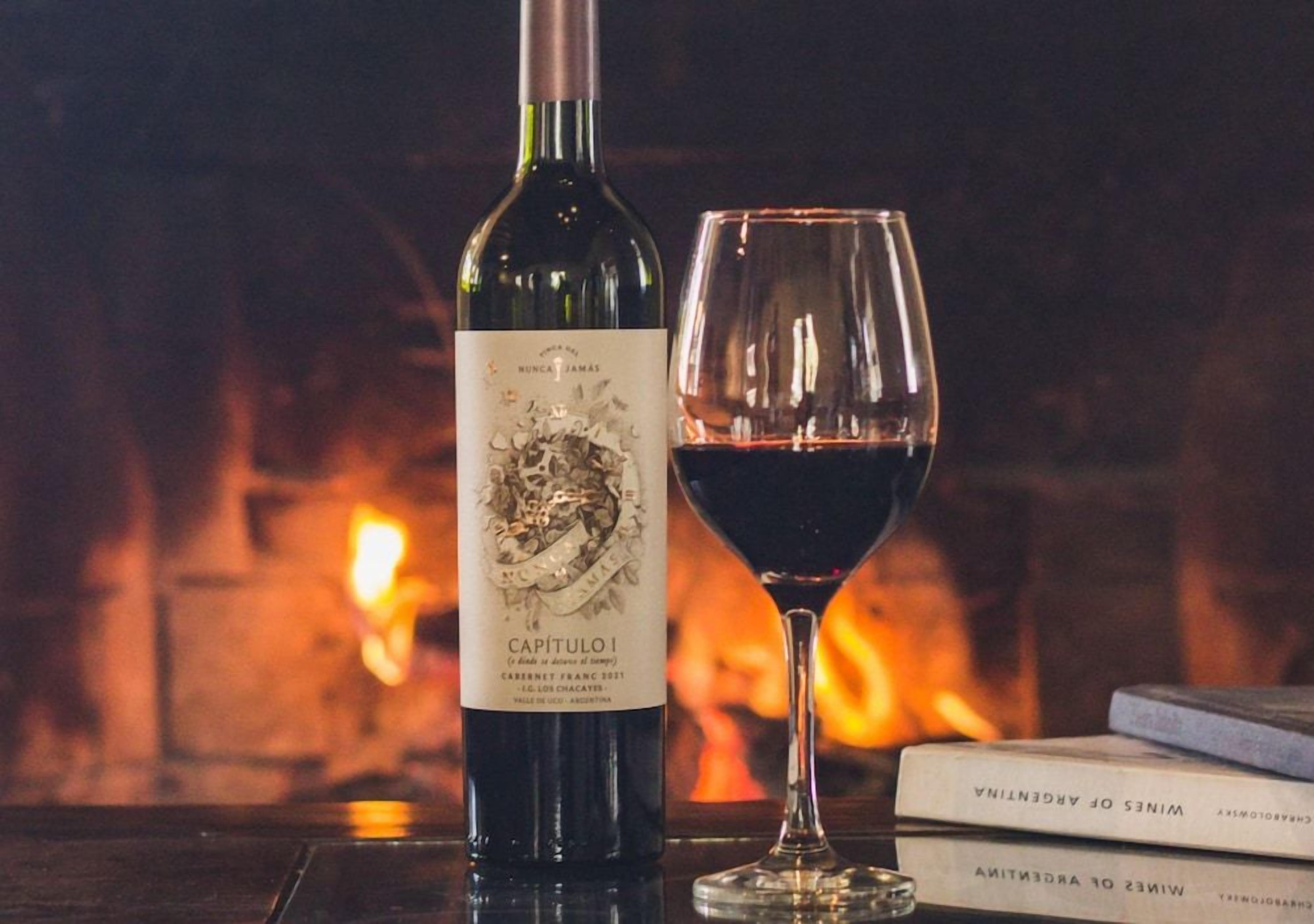 A bottle of wine and a glass of red wine on a table with a fireplace in the background.
