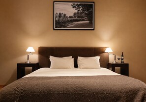 Ein Hotelzimmer mit einem großen Bett, zwei Nachttischen mit Lampen, einem gerahmten Bild an der Wand und einer Weinflasche auf einem der Tische.