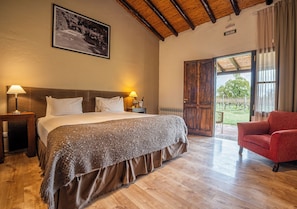 Un dormitorio con cama, lámparas de noche, una silla roja, suelo de madera, una puerta que da a un espacio exterior y un cuadro en la pared.