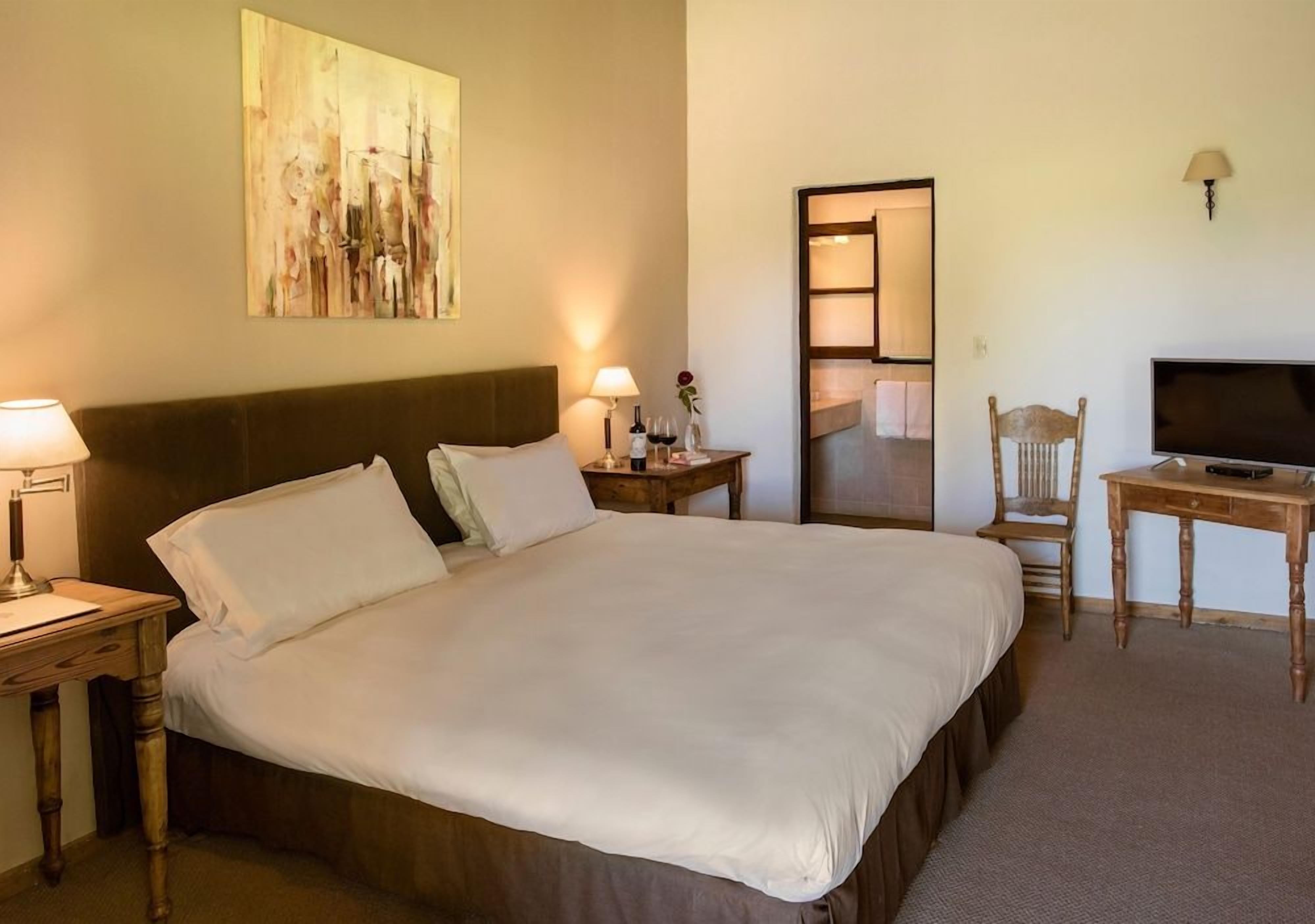 A hotel room with a large bed, a desk with a television, a chair, a bedside table with a lamp, and a painting on the wall.