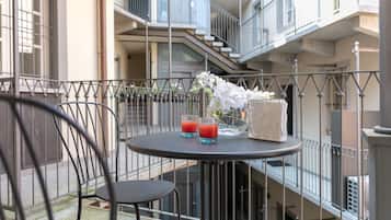 Un balcone con un tavolo rotondo, due sedie, un vaso con fiori bianchi e due bicchieri di bevanda rossa.