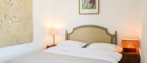 Un lit avec du linge de lit blanc, une tête de lit, une lampe de chevet et un tableau accroché au mur.