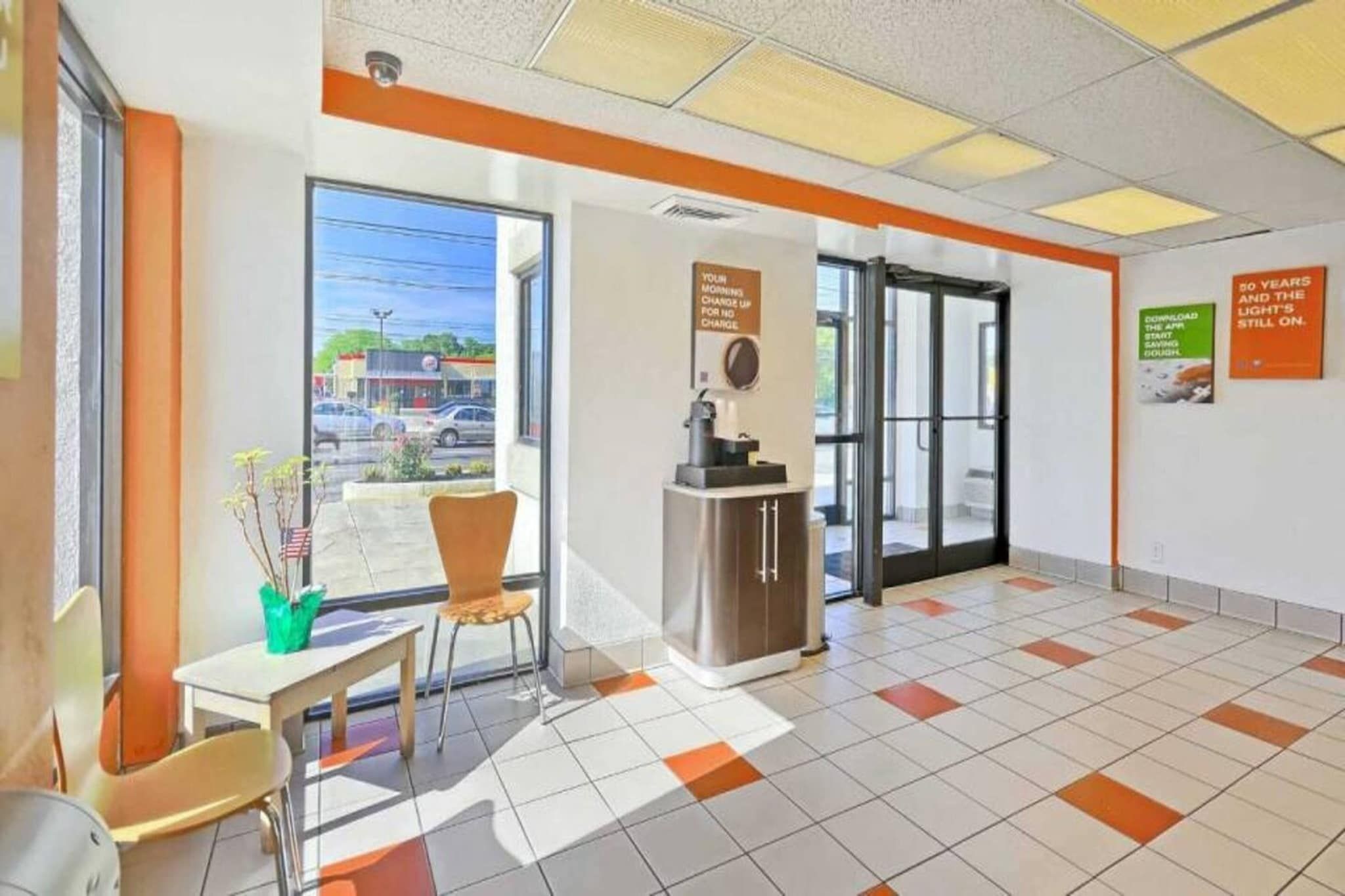 A hotel lobby with a reception desk, a seating area with chairs, a coffee machine, and a view of the street through large windows.