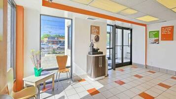 A hotel lobby with a reception desk, a seating area with chairs, a coffee machine, and a view of the street through large windows.