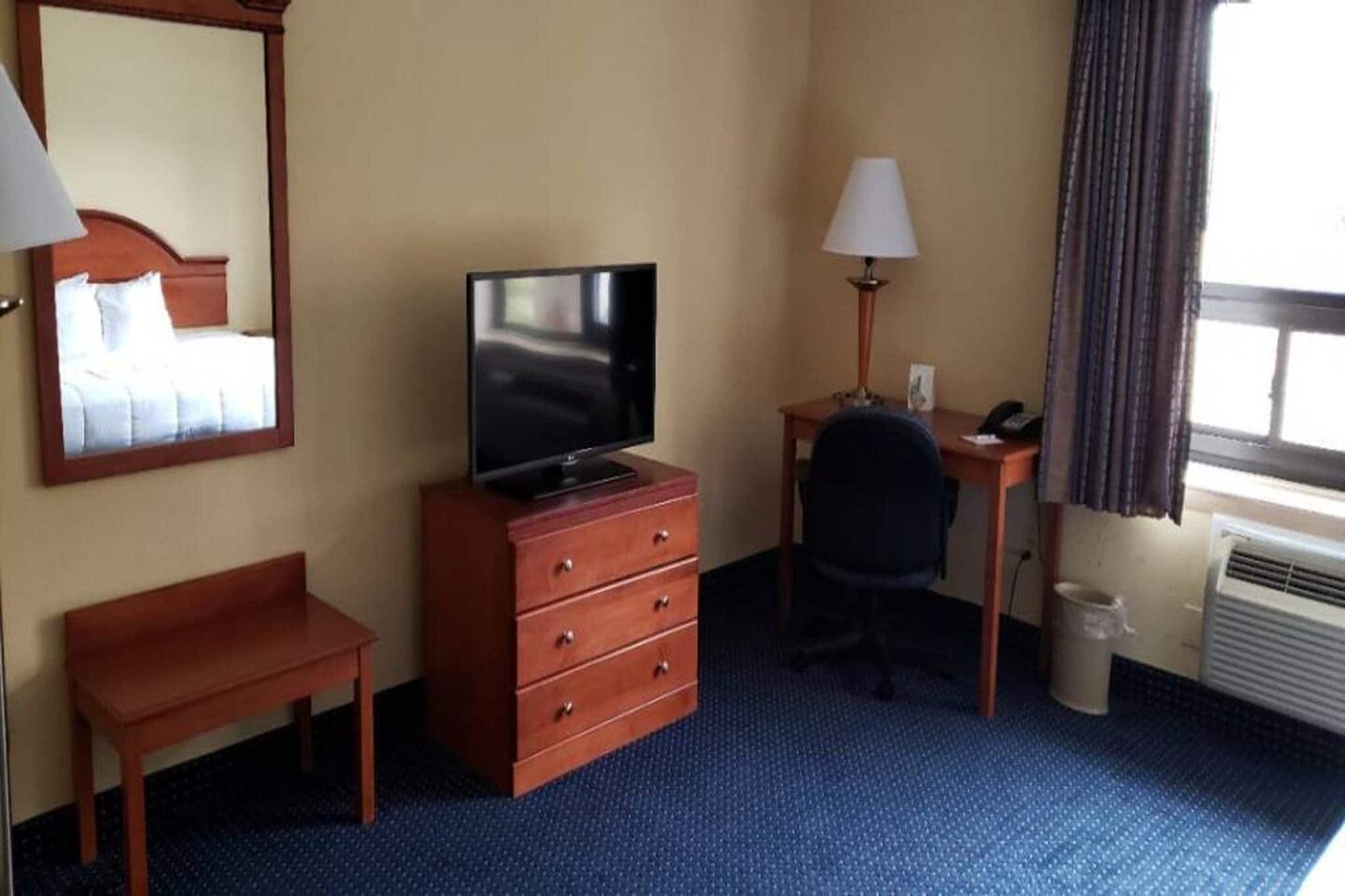 Ett hotellrum med en säng, ett skrivbord med en lampa, en tv, en stol och en papperskorg.