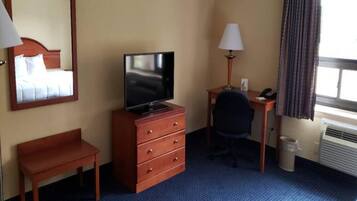 Ett hotellrum med en säng, ett skrivbord med en lampa, en tv, en stol och en papperskorg.