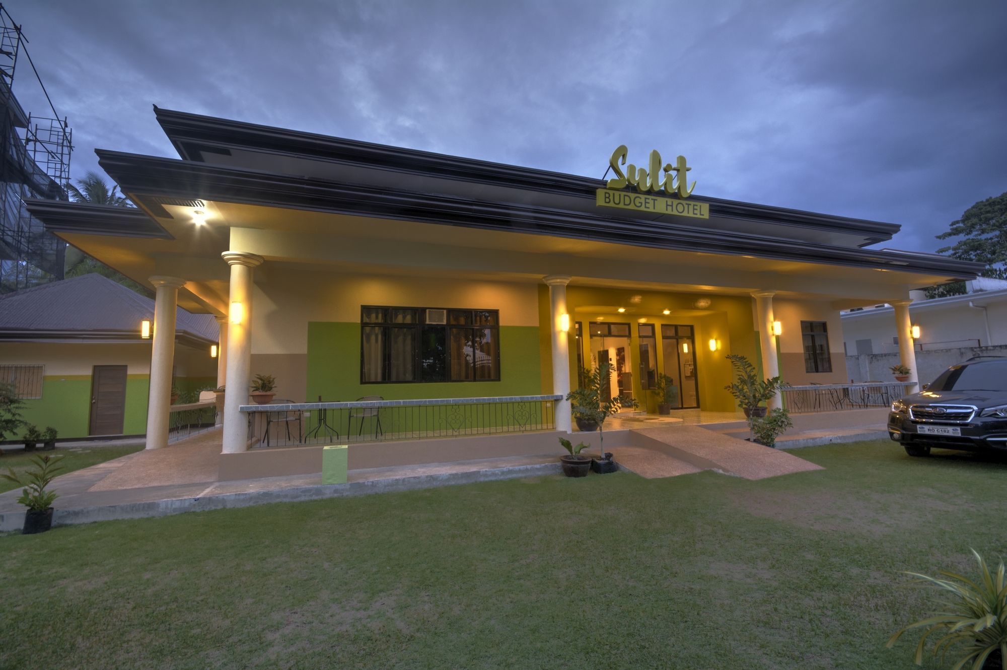 A hotel with a covered entrance, a sign reading 'Sudit Budget Hotel,' and a car parked outside.