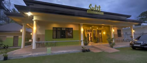 A hotel with a covered entrance, a sign reading 'Sudit Budget Hotel,' and a car parked outside.