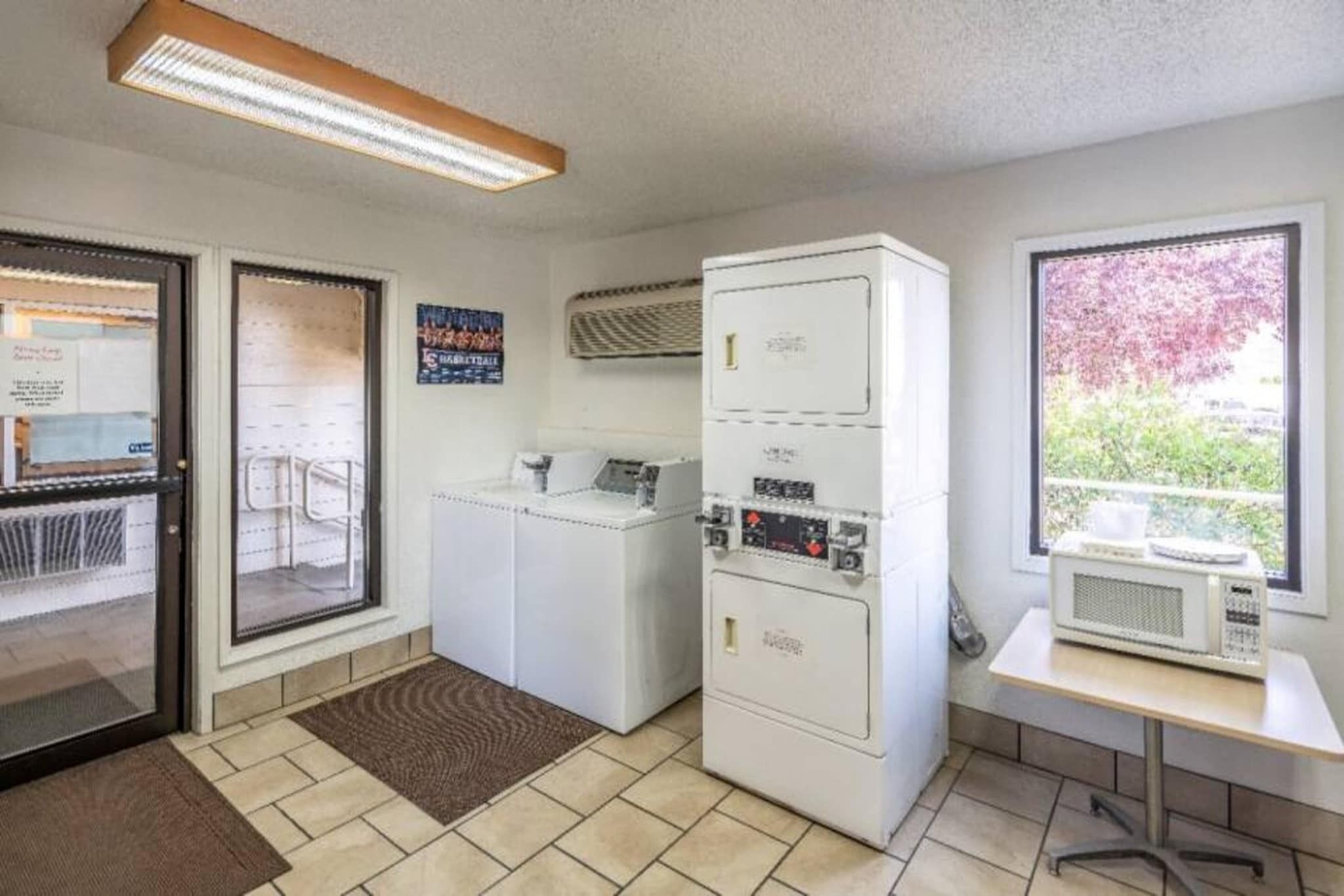 A laundry room with a washing machine, dryer, and microwave. There are two doors, one leading to a balcony with a view of trees.