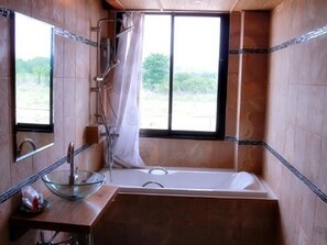 A bathroom with a bathtub, a glass bowl sink, a shower, and a window with a view of greenery.