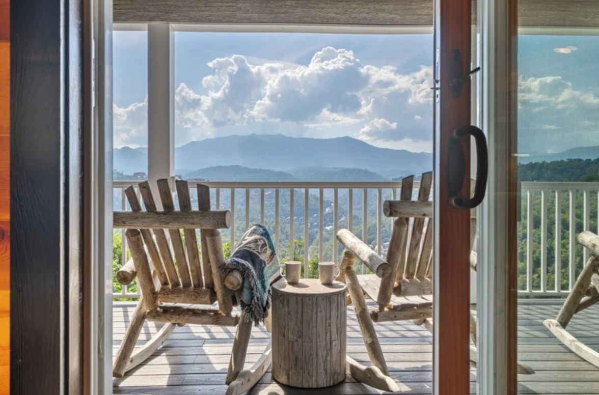 A wooden deck with two Adirondack chairs, a small round table, and a view of mountains.