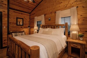 A wooden cabin bedroom with a large bed, two bedside lamps, a nightstand with a clock, and a mirror on the wall. - Smoky Mountain View | Pickleball | Family Retreat (Gatlinburg)