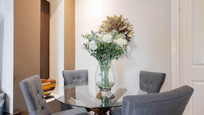 A dining area with a glass table surrounded by four grey upholstered chairs. A vase with white flowers and a decorative wall piece are on the table. - Carmel Apartments (Falkirk)