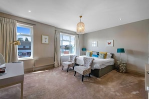A bedroom with a bed, two armchairs, a desk, a window with curtains, and a hanging light fixture.