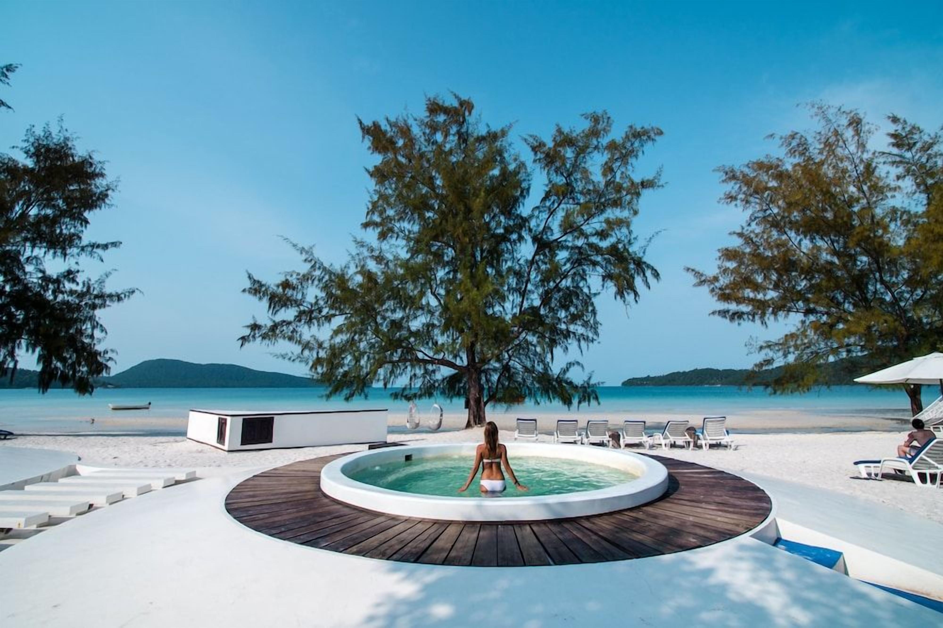 A pool area with a circular wooden deck, a person in the pool, and beach chairs and umbrellas.