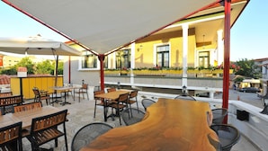 An outdoor seating area with tables and chairs, a wooden table centerpiece, and a building with yellow walls and windows in the background. - Petrino Gokceada Hotel (Gökçeada)