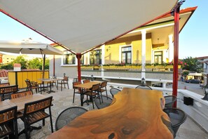 Een terras met tafels en stoelen, een houten decoratiestuk op tafel, en een gebouw met gele muren en ramen op de achtergrond.