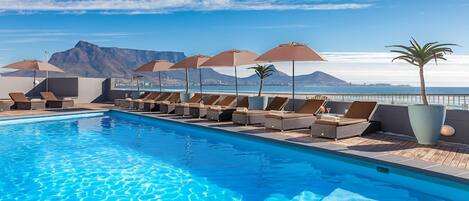 A poolside area with lounge chairs and umbrellas, a clear sky, and a mountain view.