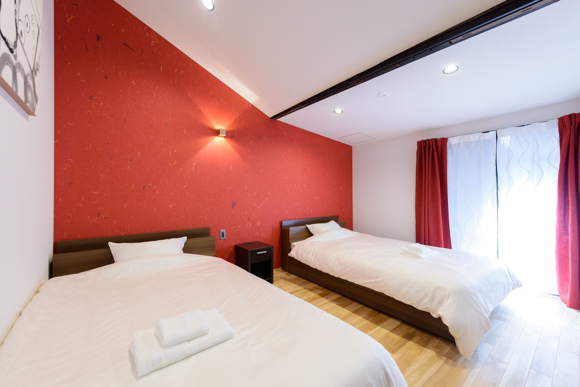 A hotel room with two beds, a red textured wall, a wooden headboard, a bedside table, and a window with red curtains.