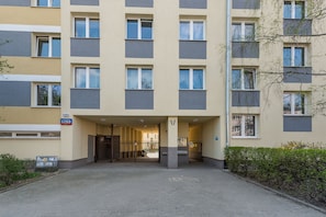 Ein mehrstöckiges Gebäude mit einem zentralen Eingang, einer Adresse auf einem Schild und einem durch ein Tor gekennzeichneten Parkbereich.