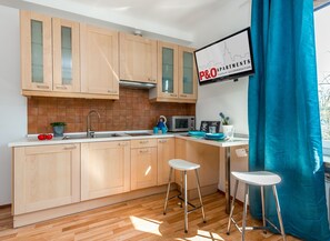 A modern kitchen with wooden cabinets, a microwave, and a sink. There are two stools and a window with a curtain. - P&O Waszyngtona (Warsaw)