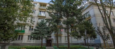 A multi-story residential building with a central entrance, surrounded by trees and a paved courtyard.