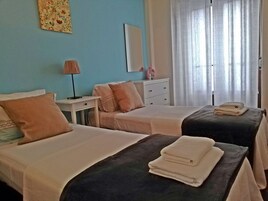 Quarto de hotel com duas camas, mesa de cabeceira com abajur, cômoda e janela com cortinas.