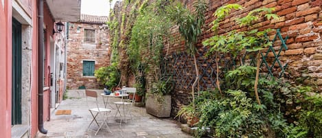 A narrow alleyway with a brick wall on one side and a building on the other. There is a small outdoor seating area with a table, chairs, and potted plants.
