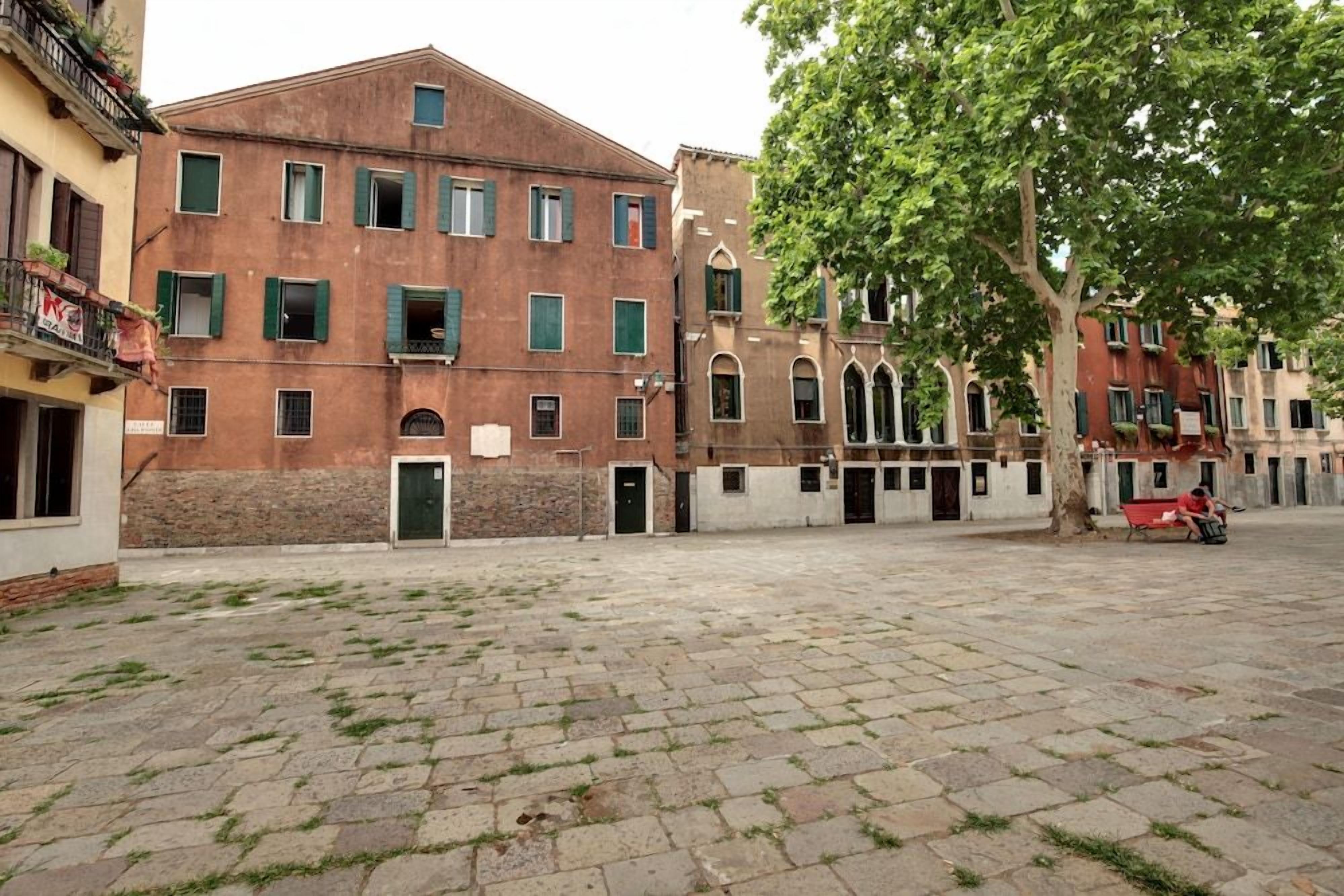 Pátio com chão pavimentado, uma árvore e um edifício com várias janelas e varandas.