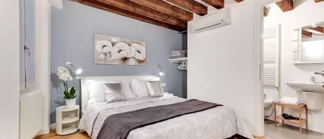 Quarto com cama, mesas de cabeceira, ar condicionado fixado na parede, radiador branco, teto de madeira e banheiro com pia e espelho.