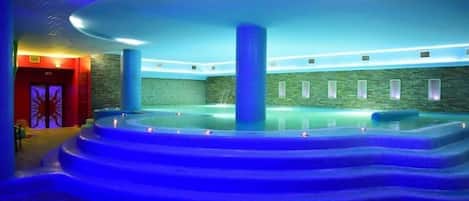 An indoor swimming pool with a blue-lit design, surrounded by steps and stone walls.