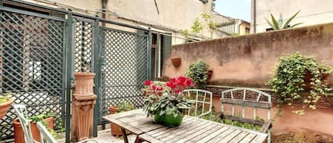 A courtyard with a wooden table and chairs, surrounded by potted plants and a metal gate.