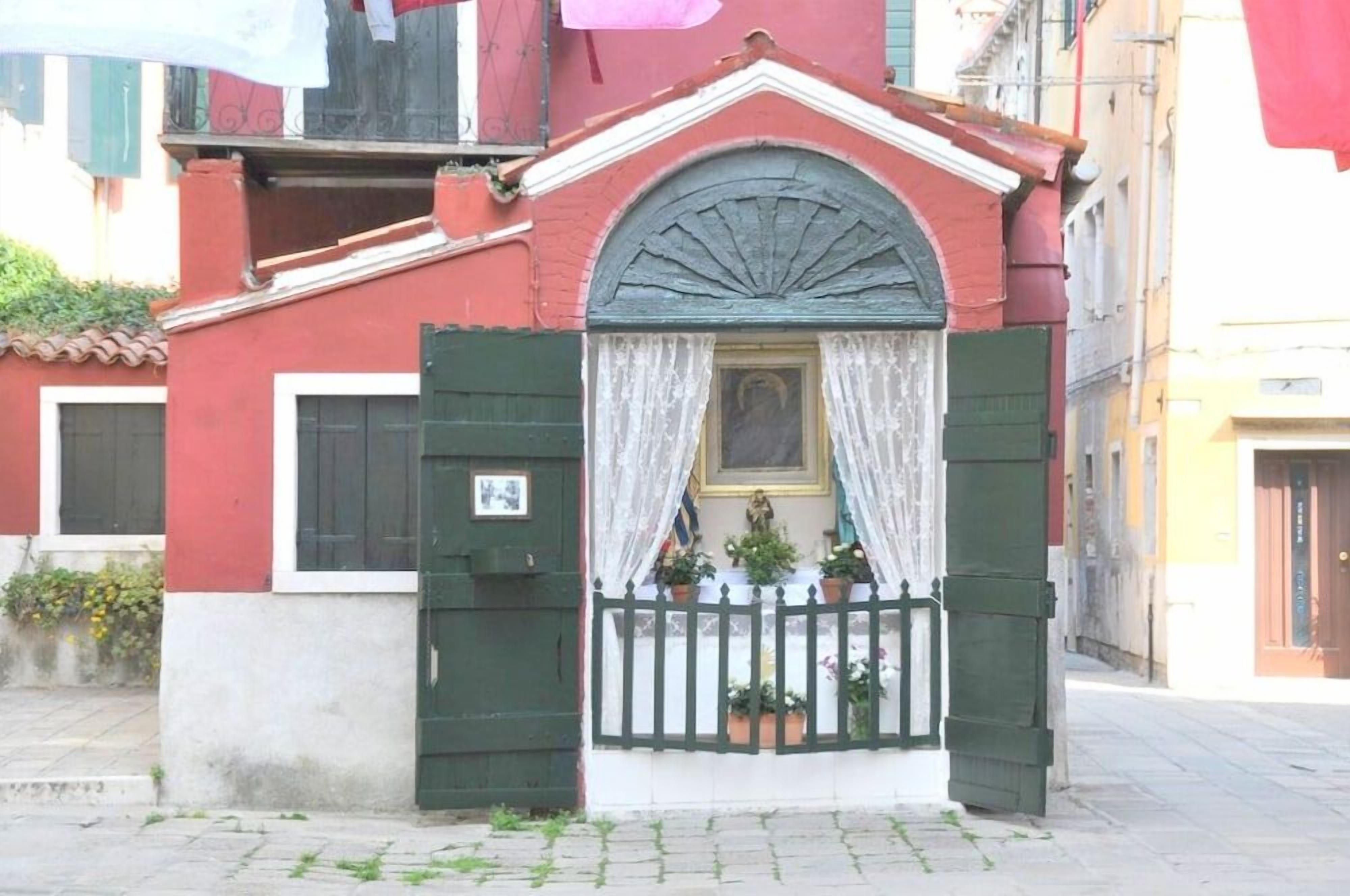 Un edificio con fachada roja, una puerta blanca con marco verde y un pequeño balcón con plantas en macetas.