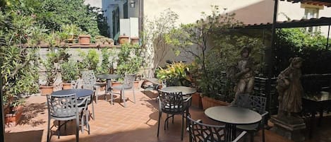 Un patio al aire libre con varias mesas y sillas, rodeado de plantas en macetas y estatuas.
