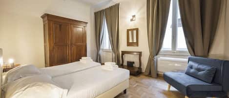 Quarto de hotel com cama, sofá, guarda-roupa de madeira, mesa pequena e janela com cortinas.