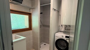 Ein kleines Badezimmer mit Dusche, Waschmaschine und einem Fenster mit Jalousien.
