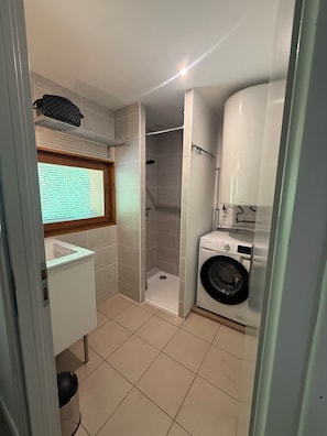 Een kleine badkamer met een douche, een wasmachine en een raam met zonwering.