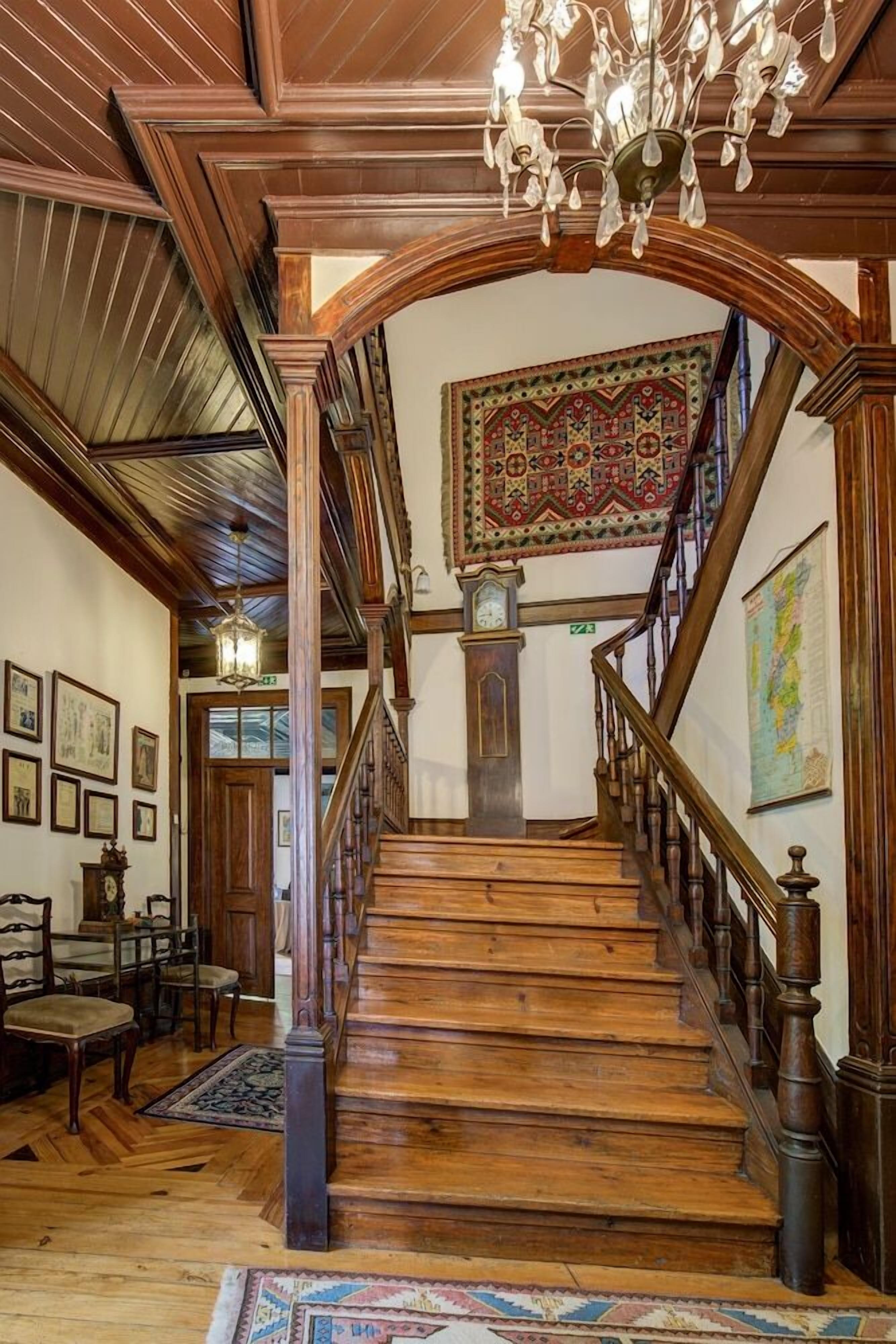 A grand staircase with wooden railings and a chandelier, leading to a room with a large patterned rug and framed pictures on the walls.