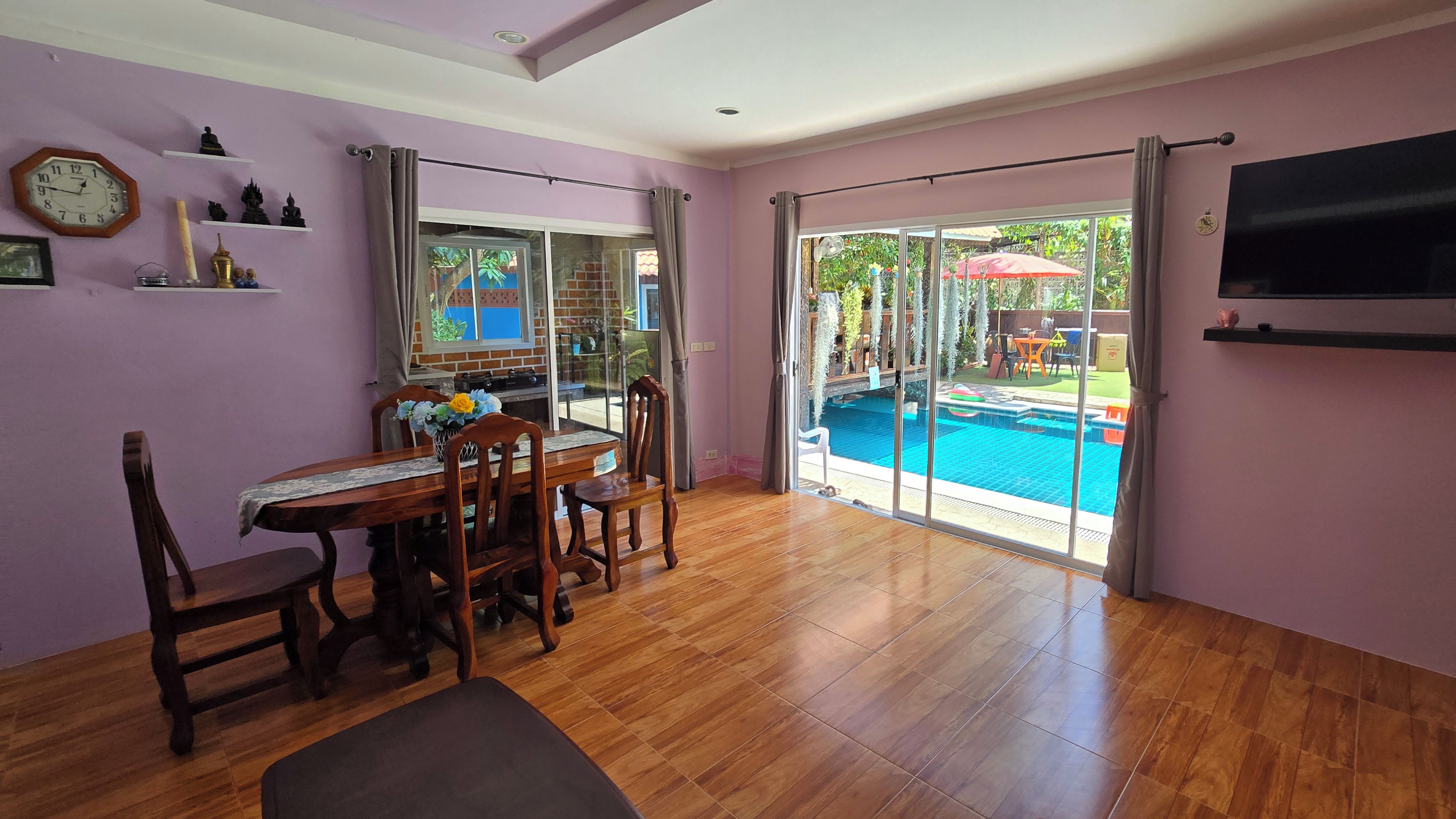 A room with a dining table, chairs, a TV mounted on the wall, and a sliding glass door leading to an outdoor swimming pool area.