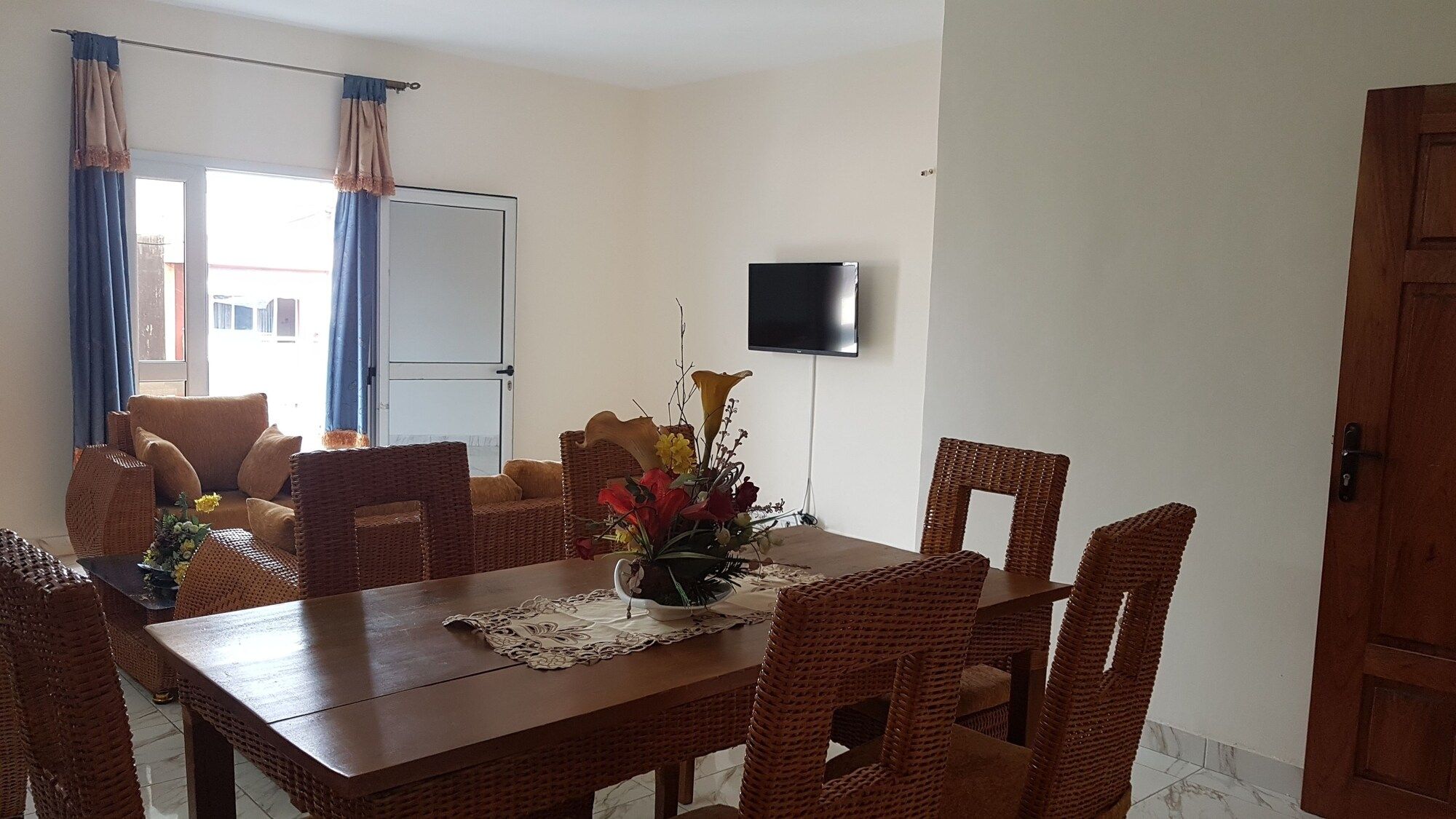 Un coin repas avec une table en bois et des chaises en osier, un vase rempli de fleurs et un téléviseur fixé au mur.