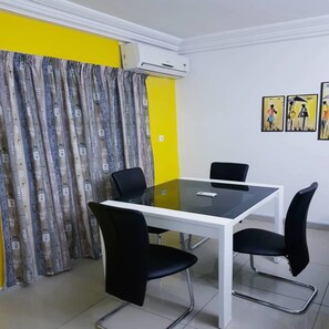 Une salle de réunion avec une table en verre, quatre chaises noires, un pied de table blanc, un mur jaune, un rideau gris et un appareil de climatisation.